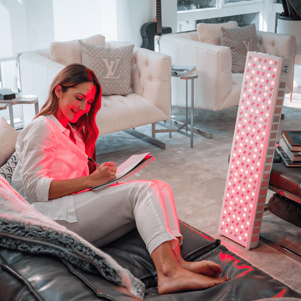 A woman sits on a couch, jotting in her notebook, as the Vital Elite Red Light therapy panel casts a warm glow throughout the modern living room.