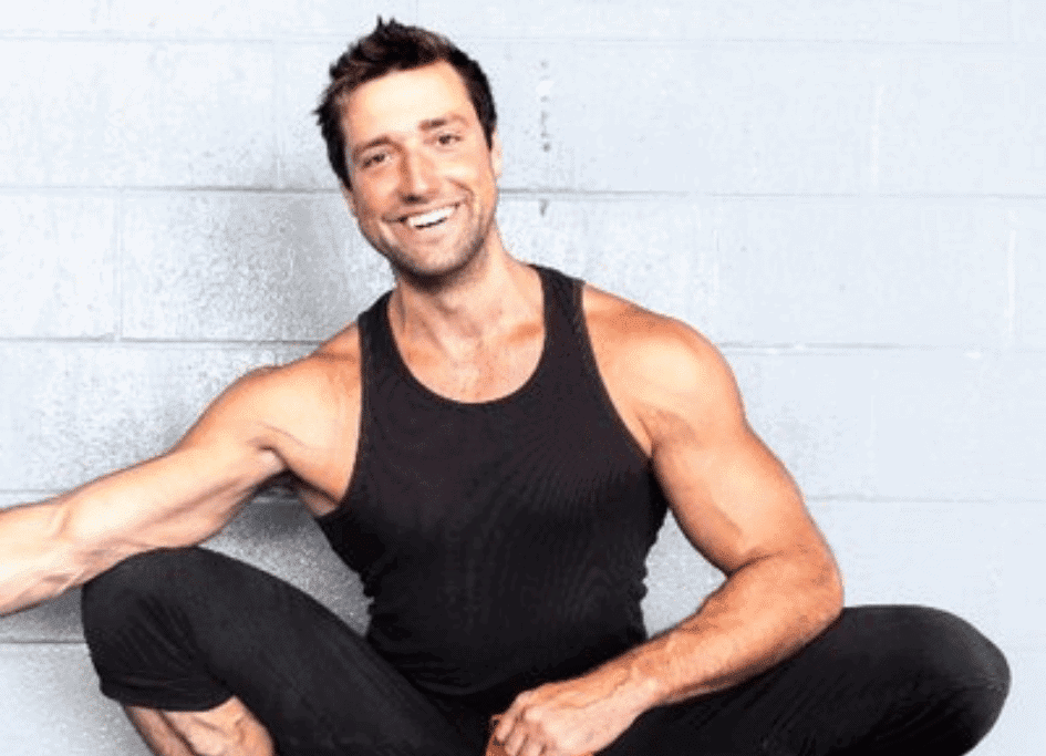 Smiling man in a black tank top crouches against a light gray wall, extending one arm forward, showcasing his dedication to the red light therapy community.