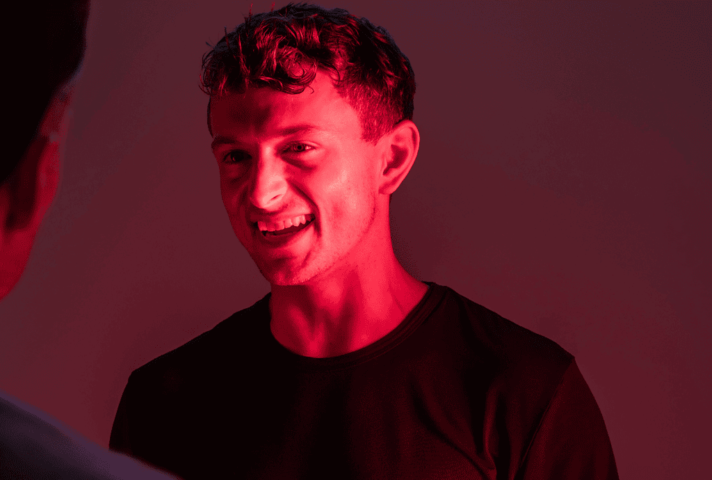 A person smiling under red lighting, donning a dark shirt, faces another who is partially visible, capturing the essence of connection. This moment could be part of an "about us" section, illustrating the warmth and camaraderie within our team.