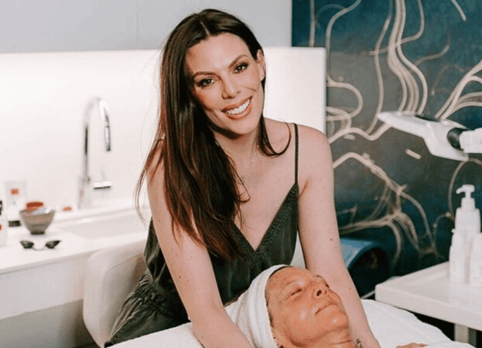 A woman stands smiling, applying a facial treatment to a person lying down in a spa setting.