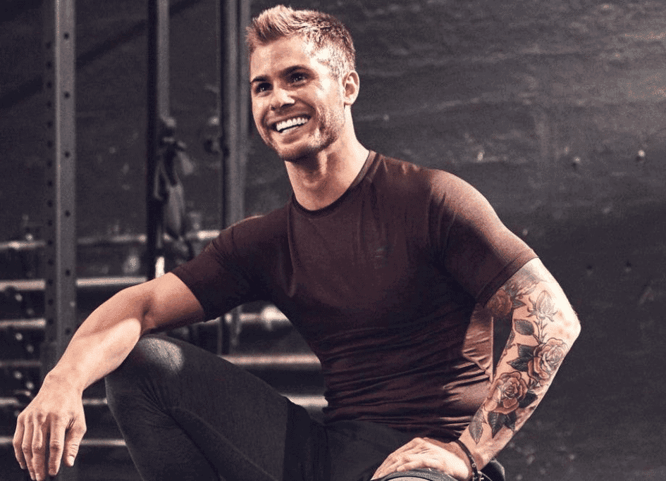 A person with a floral tattoo on their arm is smiling while sitting in a gym, wearing a dark shirt and pants.