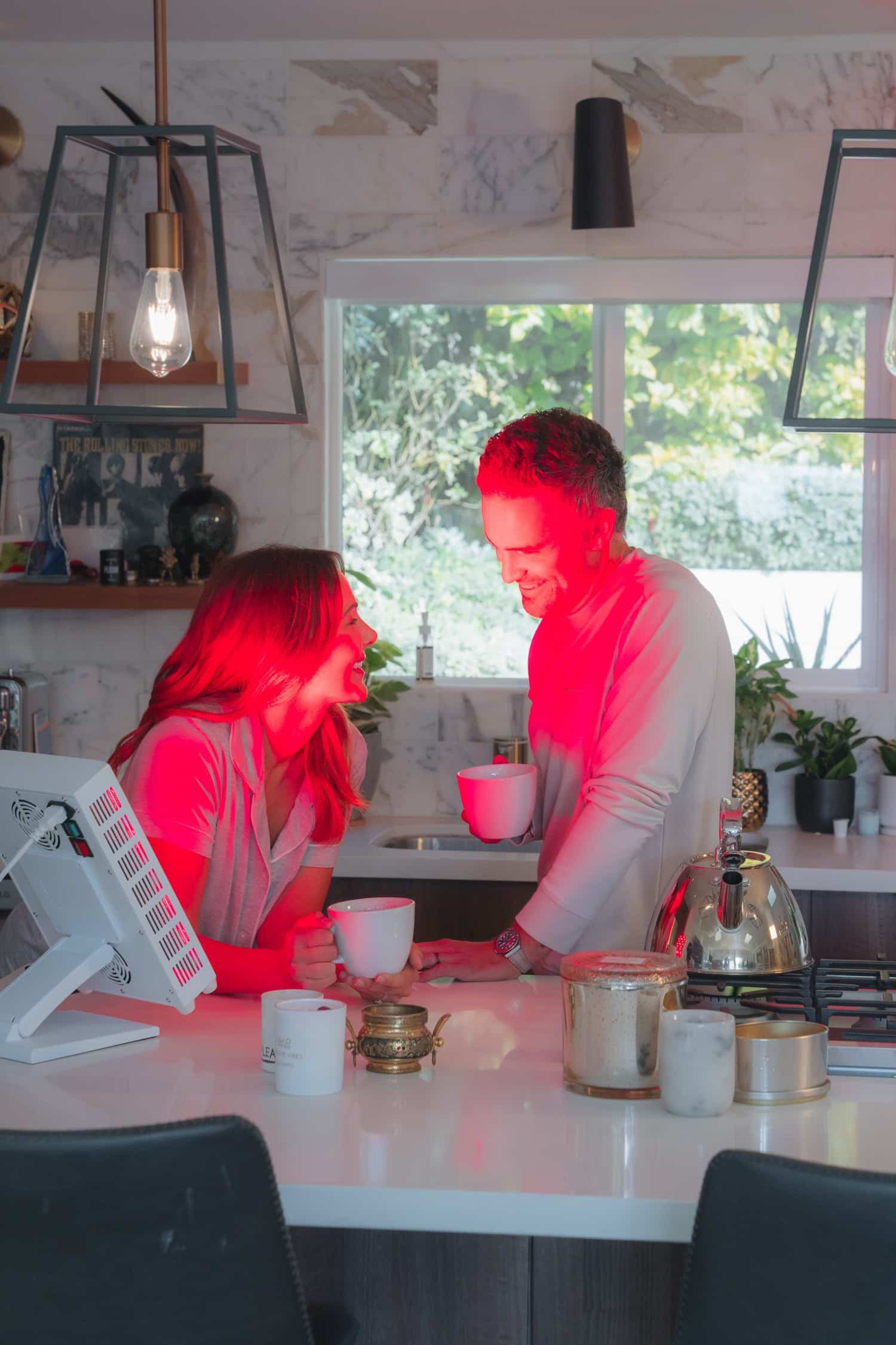 A man and woman stand in a kitchen lit by red light, each holding a mug, smiling at each other across a counter.
