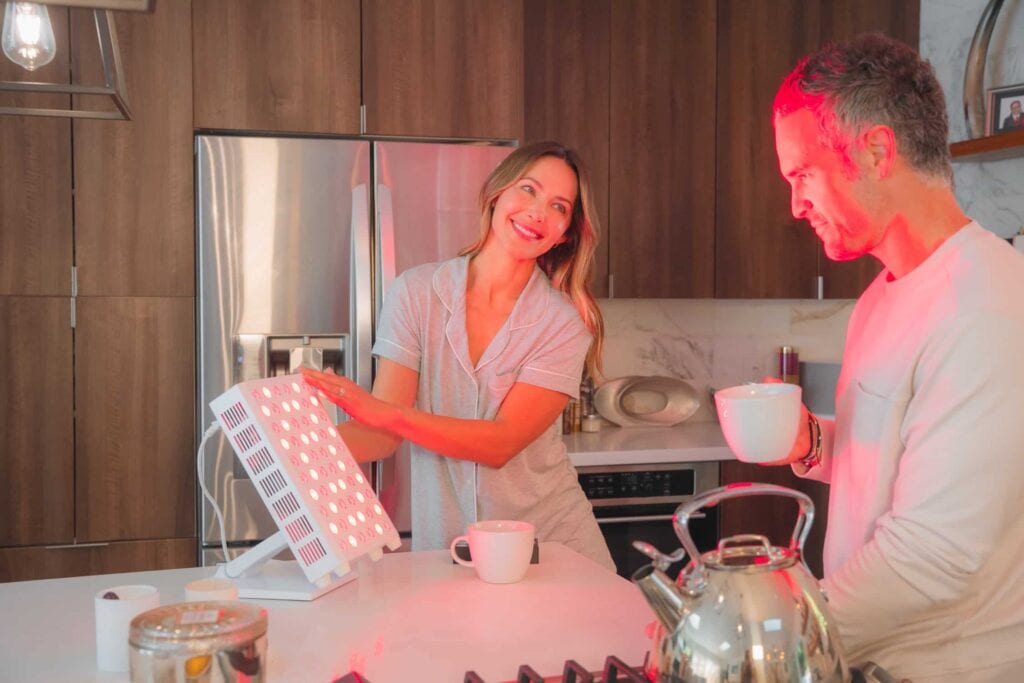 Two people in a kitchen use a red light therapy device. One operates the device while the other holds a mug, both standing near a counter with a kettle in the foreground.