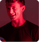 A person with short curly hair, wearing a dark shirt, smiles in red lighting.