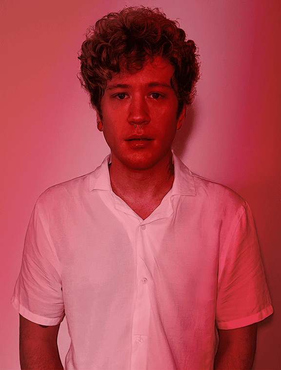 A person with curly hair in a white shirt stands confidently against a gradient red backdrop, looking directly at the camera. This image perfectly captures the essence of our "About Us" spirit—bold, direct, and unmistakably vibrant.