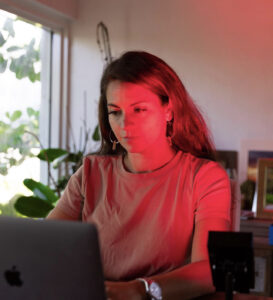 A woman in a pink shirt focuses on her laptop, analyzing a dashboard, while red light therapy for acne illuminates the room. Nearby, plants thrive by the window, creating a serene workspace.
