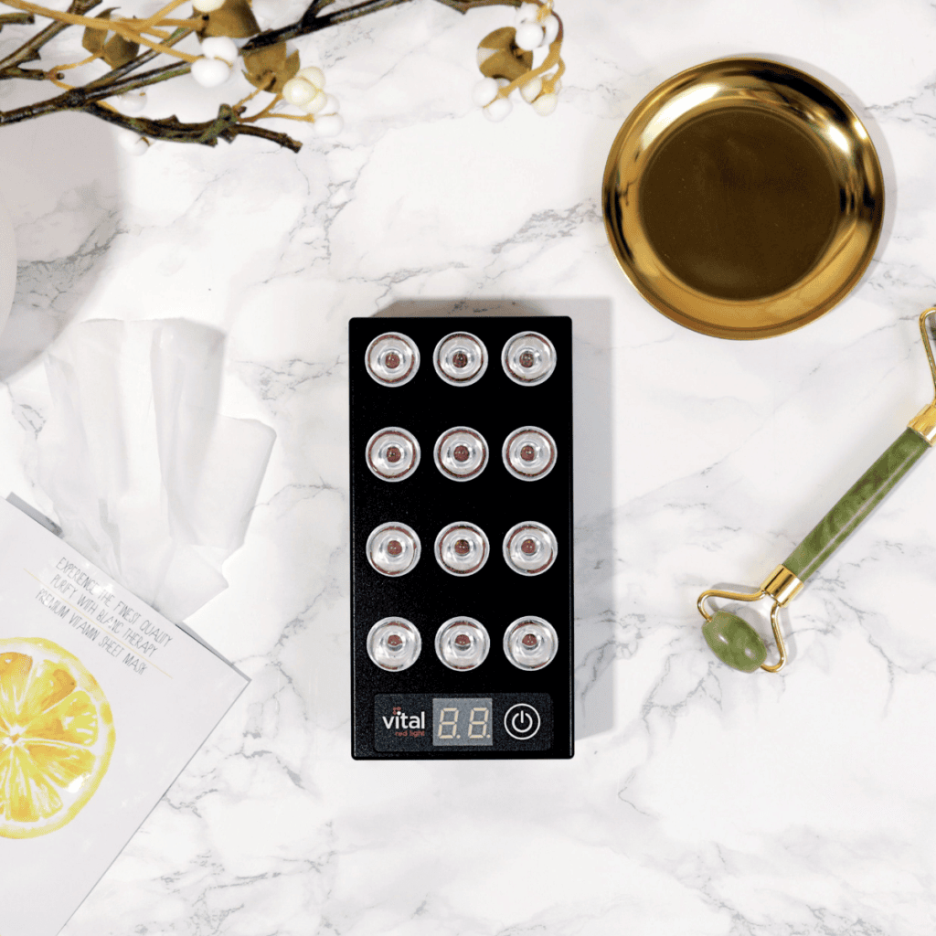 Flat lay of a skincare setup on a marble dashboard. Central black device with multiple circular lights, a jade roller, a golden tray, tissues, and a card with a lemon slice design. Perfect for wholesale beauty enthusiasts seeking elegant organization.