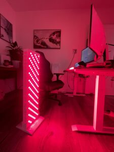 A room lit with pink light, featuring a standing desk and computer monitor displaying a vibrant dashboard, alongside an office chair. A large vertical LED light panel casts a warm red glow over this dynamic workspace.
