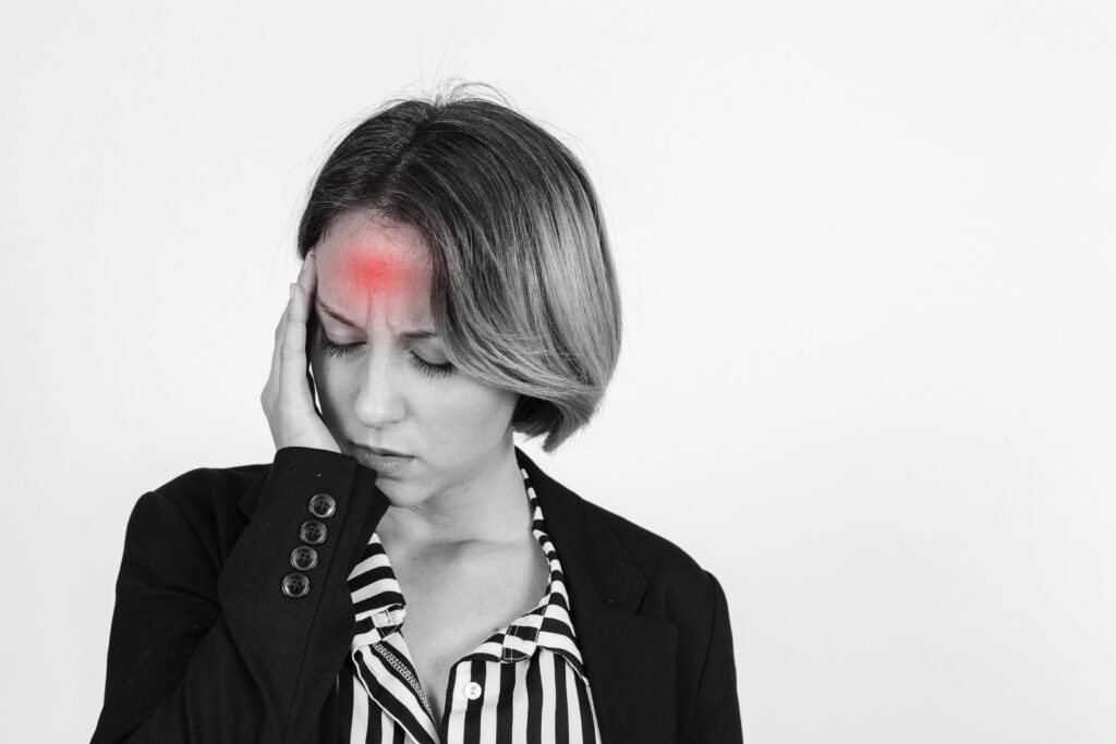 A woman with short hair holds her head, showing signs of a headache. A red highlight marks her forehead, suggesting pain. She wears a black blazer and a striped shirt, considering red light therapy for concussion relief.