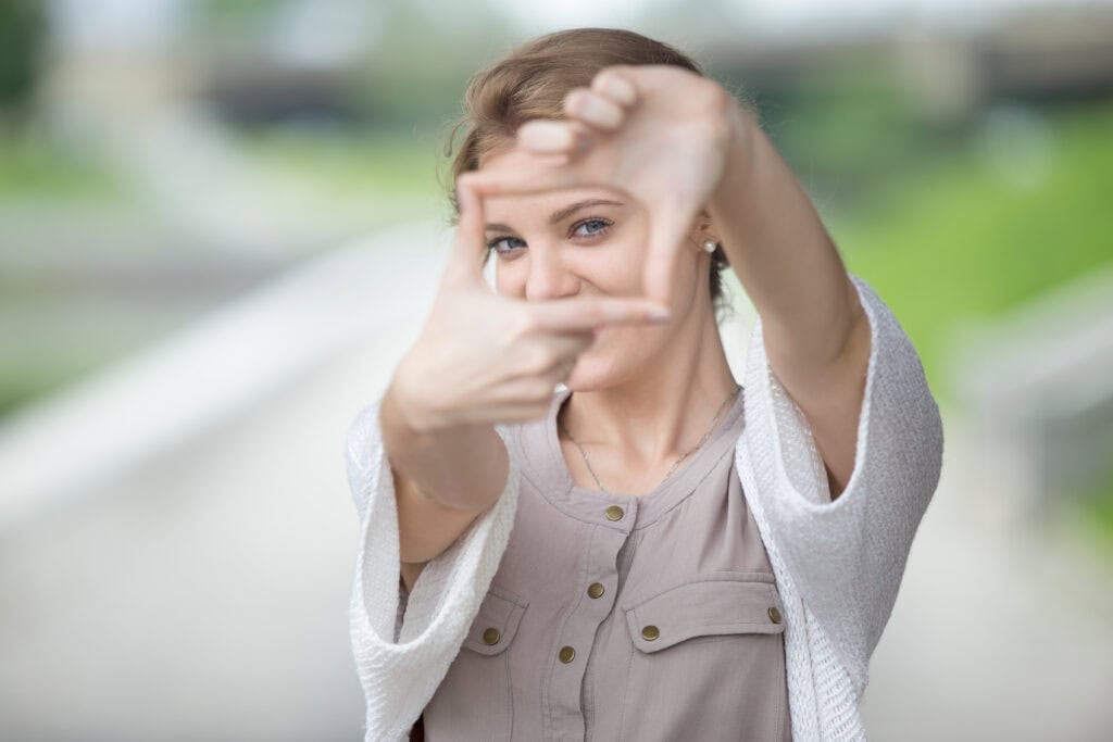 A woman in a beige blouse and white cardigan frames her face with her fingers, smiling outdoors, while raising awareness about age-related diseases like macular degeneration.