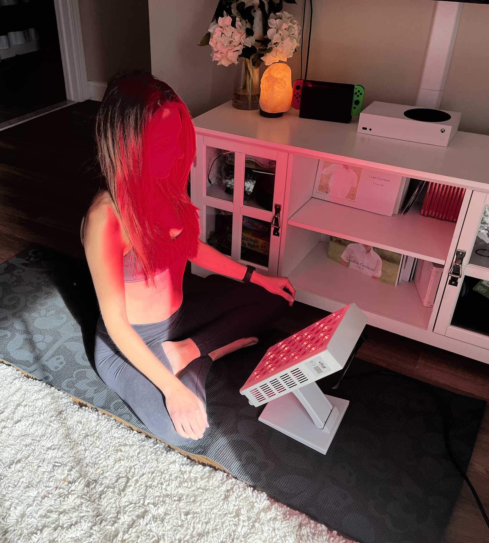 A person sitting cross-legged on a yoga mat, facing a red light therapy device. Nearby are shelves with a few electronic devices and decorative items.