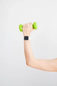 A hand wearing a smartwatch holds a small green dumbbell against a plain white background biohacking health