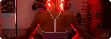 A person is sitting cross-legged, illuminated by the glow of infrared lights in a dimly lit room filled with fitness equipment, creating a serene atmosphere—a building dedicated to wellness and rejuvenation.