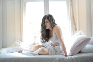 Woman sitting on a bed, holding her stomach and appearing to be in discomfort. Bright light from a large window illuminates the room. Chronic pain and sleep