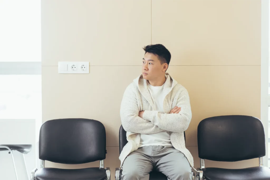 Man sitting with arms crossed on an empty chair between two vacant ones, exuding an air of serene gratification in the quiet room with beige walls, as he contemplates the fleeting nature of instant gratification.
